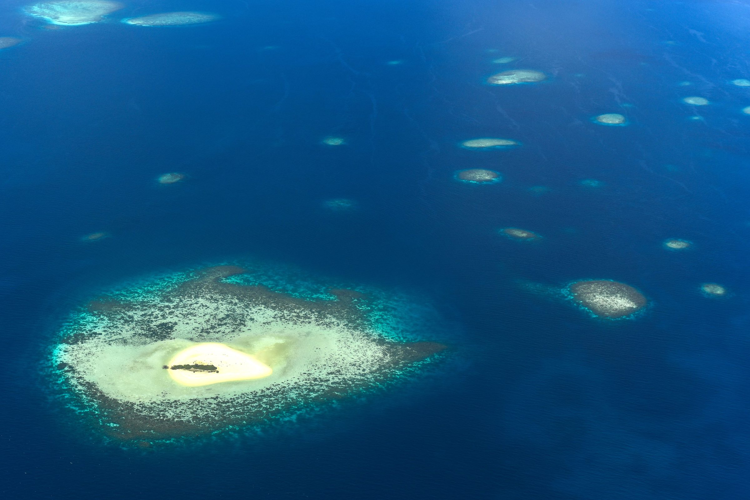 Un pequeño cayo de arena rodeado de arrecife de coral en aguas azules abiertas, visto desde el aire